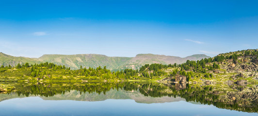 Majestic panoramic landscape at Blue montain lake in front of mountain range covered with pine forest and reflection of clear sky in water in wild taiga, Siberia, Russia