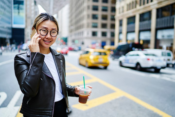 Happy female with takeaway beverage having international contact phoning for discussing mobility connection, pretty Asian woman in leather jacket phoning via cellphone application and 4g roaming