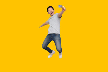 Asian man jumping and showing credit card on hand isolated on yellow background