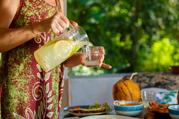 healthy food and Breakfast concept - female hands pouring lemonade from the decanter into glass on the Villa's terrace , glass pitcher with slices of lemon, lime, water, mint leaves, selective focus