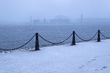 Winter view during a heavy snowfall in St. Petersburg, Russia. White snow cover and fence of the river embankment in the form of pillars and chains.