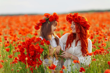 Little girl with redhead mother in white dresses and wreathes poses with bouquet of poppies on poppy field at summer sunset