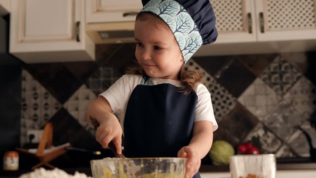 Cute little smiling girl in apron and chef hat is kneading the dough. The child is all dirty with flour. Slow motion.