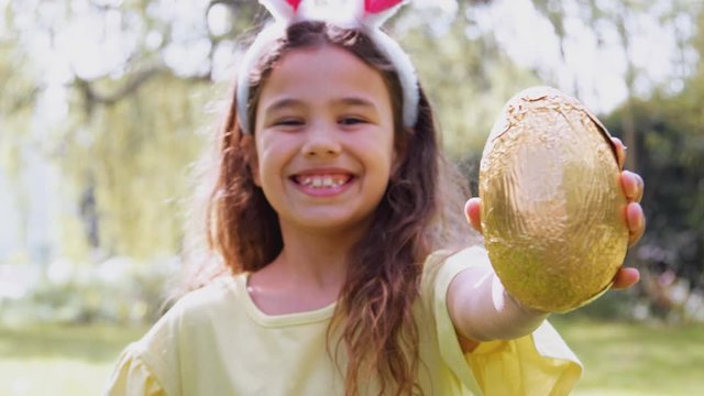 Portrait Of Girl Wearing Bunny Ears Holding Chocolate Egg On Easter Egg Hunt In Garden