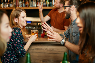 Handsome caucasian man with eyeglasses hugging his girlfriend and talking to her while standing in a pub and drinking beer. Nightlife.