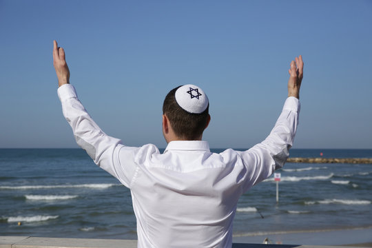 young Jew man in a kip pah hat is standing with his back with raised hands praying against the blue sea and blue sky.