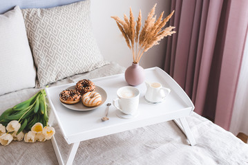 Breakfast in bed, cup with cappuccino, doughnuts, flowers white tulips, morning concept, linen bed sheets, woman's day