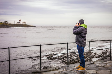 Young man with camera taking pictures of lighthouse by the sea far away in cold day