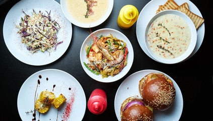 Assorted different dishes on a black background. Dining table with burgers, cream soups, wok noodles, cole slaw salad and grilled corn. Top view, food flat lay