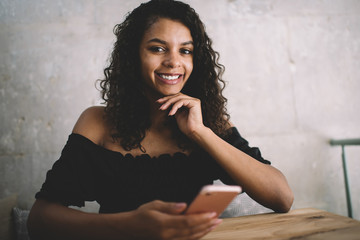 Cheerful young ethnic woman using smartphone while chilling in light modern cafe