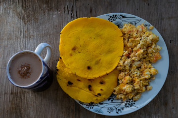 Dos arepas de maiz amarillo, con huevo revuelto en un lado, en un plato blanco decorado con flores y una taza de chocolate con leche, sobre madera rustica 