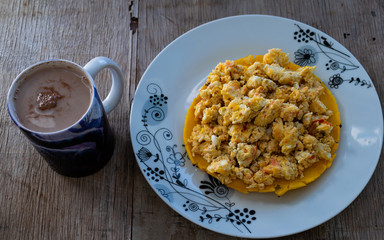 arepa de maiz amarillo, con huevo revuelto hecho con cebolla y tomate, encima, en un plato blanco, con una taza de chocolate con leche en el lado izquierdo 