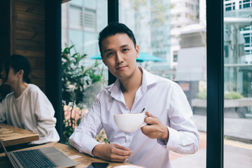 Young Asian man with coffee cup sitting at table with laptop