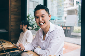 Asian smiling young male with smartphone sitting at desk and looking at camera