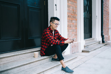 Pleased young man in checked shirt browsing smartphone on doorstep