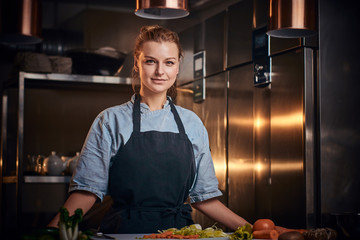 Confident and serious female chef standing in a dark kitchen next to cutting board with vegetables...