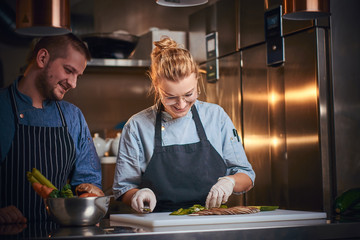 Smiling male and female chefs standing in a dark kitchen next to cutting board with vegetables on...