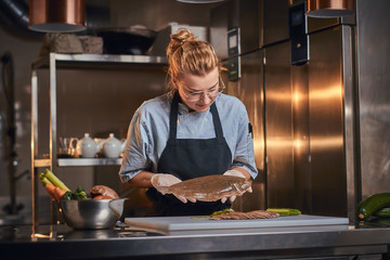 Beautiful and stylish woman chef standing in a dark restaurant kitchen next to cutting board with vegetables on it, holding a big fish and wearing apron and denim shirt, posing for the camera, cooking