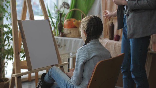 Teacher at art Studio teaches children, the girl sitting in front of his easel carefully listens to the teacher.