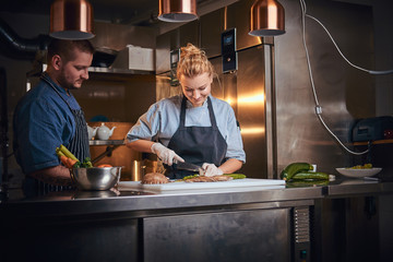 Serious male and female chefs standing in a dark kitchen next to cutting board with vegetables on it, wearing aprons and denim shirts, posing for the camera, cooking show style, working together