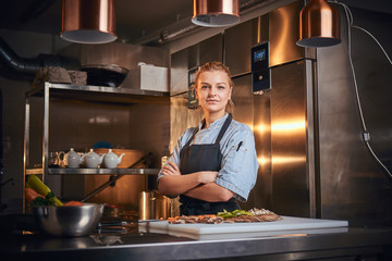 Confident and serious female chef standing with hands crossed in a dark kitchen next to cutting board with vegetables on it, wearing apron and denim shirt, looking in the camera, cooking show look - Powered by Adobe