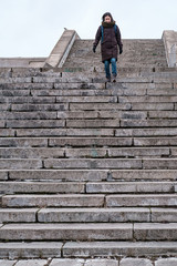 Satisfied tourist in warm clothes and with a city backpack goes down the old stone steps of a dangerous staircase. Bottom view.