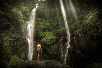Young lonely man traveler exploring Bali waterfall in tropical forest in mountains as travel lifestyle