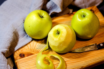 Three fresh green apples lie on a wooden cutting Board, along with a gray linen napkin and a knife. One of the apples has been peeled off. Selective focus. Close up.