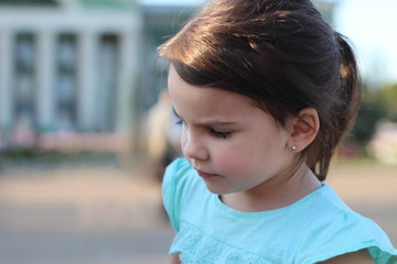 Beautiful little preschooler girl with dark hair and a ponytail on the background of a blurry summer building