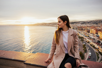 Fototapeta na wymiar Young beautiful woman admiring panoramic view of european street in Nice, France on sunset. Architecture high from the balcony. Girl looking at street and sea panorama on sunset. 