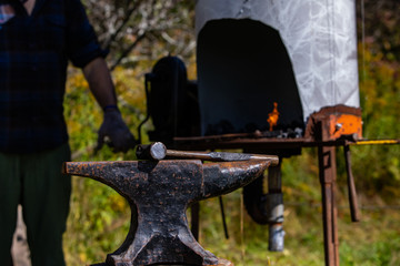 A close up selective focus shot of a metalsmith anvil and mallet, with blurry blacksmith and furnace in background during an earth festival, copy space to top