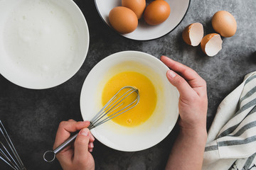 Woman's hands whisk eggs in bow. Cooking omlette for breakfast. Top view, flat lay of cooking process. Dark background.