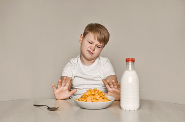The boy sits at the table and doesn 't want to eat breakfast. Tired of useful food. Moves away from himself a plate of flakes and milk.