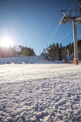 A beautiful sunny day on the mountain, the sun glows with a six-seat ski lift. Below the cable car there is a ski slope and skis and a pine forest around.
