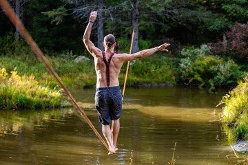 A shirtless man is seen wearing shorts and braces practicing slacklining over a woodland pond, balance, risk and strength in nature with selective focus