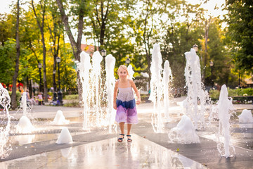 Blonde happy child girl in long purple dress running between water flow in city summer park. Child...