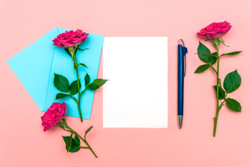 Valentine's day or other holiday concept. Mock-up of blank white message, envelope and classic blue color pen with rose flowers lying on soft pink background. Flat lay, top view