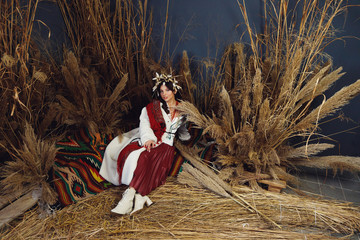 Front view of woman, wearing in national clothes and beautiful headdress, sitting on patttern colorful carpet among wheats and hayloft, looking at camera.