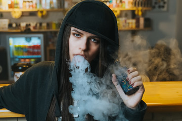 Young pretty woman in cap smoke an electronic cigarette at the vape shop. Hip-hop style. Closeup.