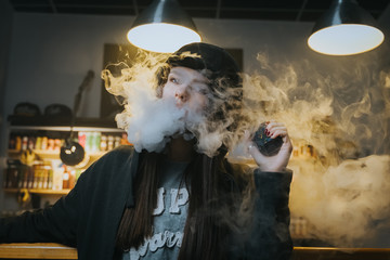 Young pretty woman in cap smoke an electronic cigarette at the vape shop. Hip-hop style. Closeup.