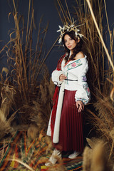 Front view of beautiful model, wearing in national clothes and chaplet with cotton and wheat on head, posing in studio among hayloft, ooking at camera.