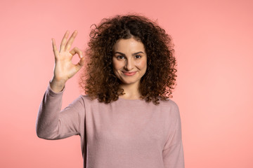 Winner. Success. Positive girl making OK sign over pink background and smiles to camera. Body language. Young curly woman with trendy glitter freckles make-up