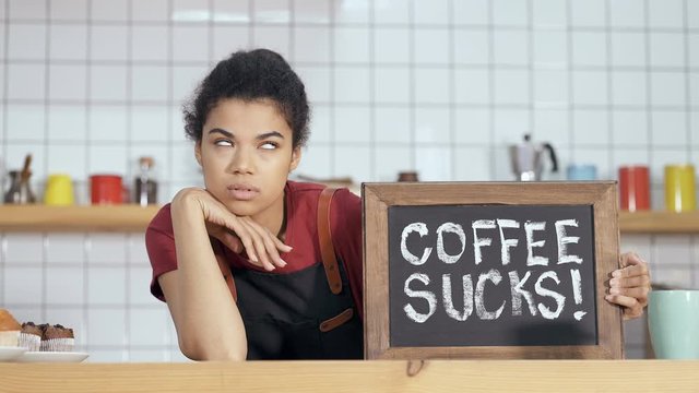 Unhappy barista holding borad with sign "Coffee sucks". Sarkasm, irony idea. Handheld shot.