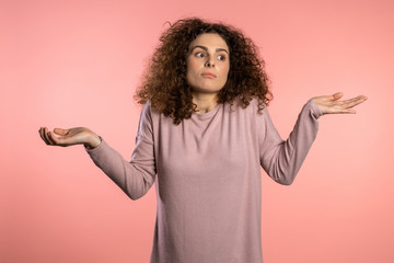  Young curly unsure girl shrugs her arms, makes gesture of I don't know, care, can't help anything...