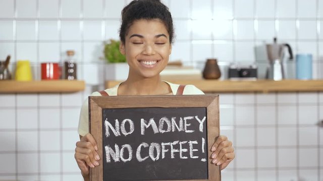 Irony and sarkasm emotion of barista worker. Standing in the coffee shop near the counter, holding board with sign "No money no coffee". Handheld shot.