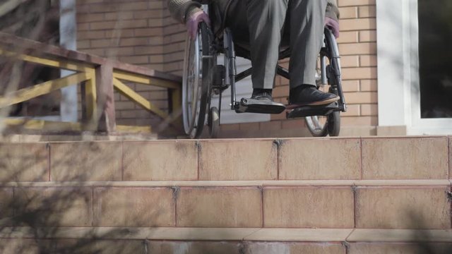 Unrecognizable elderly Caucasian man in wheelchair rolling to stairs and stopping. Disabled person having no opportunity to roll down. Ramp availability, disability problems.