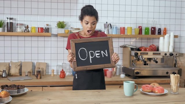 Happy, joyfull coffee shop worker holding balckboard with sign "Open" on it. Showing it in to the camera. Positive, happy and funny emotions.