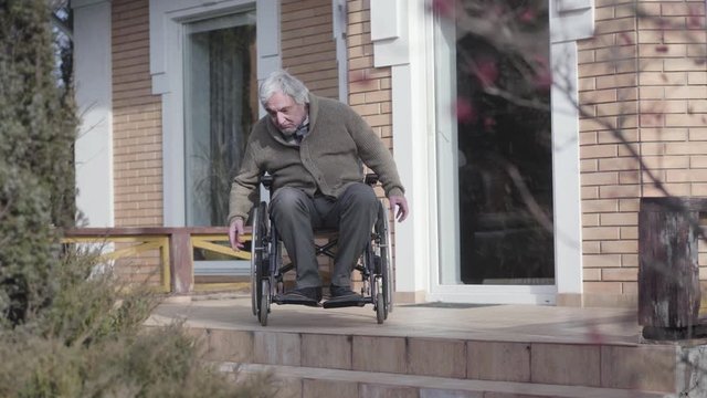 Old Caucasian man in wheelchair rolling to stairs without ramp outdoors and throwing up hands in frustration. Disabled retiree having no motion availability. Concept of disability, problem, challenge.