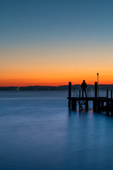 Fototapeta na wymiar A man silhouette standing on wooden pier lonely at the sea with beautifulsunset. lsunset seascape at a wooden jetty