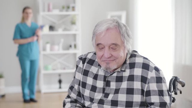 Portrait of happy Caucasian grey-haired retiree looking back at nurse, turning to camera and gesturing yes by shaking head. Old ill man living in nursing home. Help, assistance, support, health care.